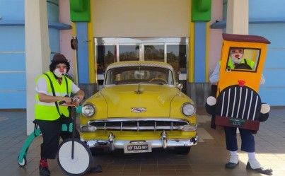 Os Palhaços Acerola e Gaguelho estão ao lado de um carro antigo amarelo, Gaguelho está de bicicleta verde com colete verde florescente e calça preta. Acerola está dentro de um caminhãozinho de espuma pintado de amarelo e preto, ele também veste um colete amarelo florescente.