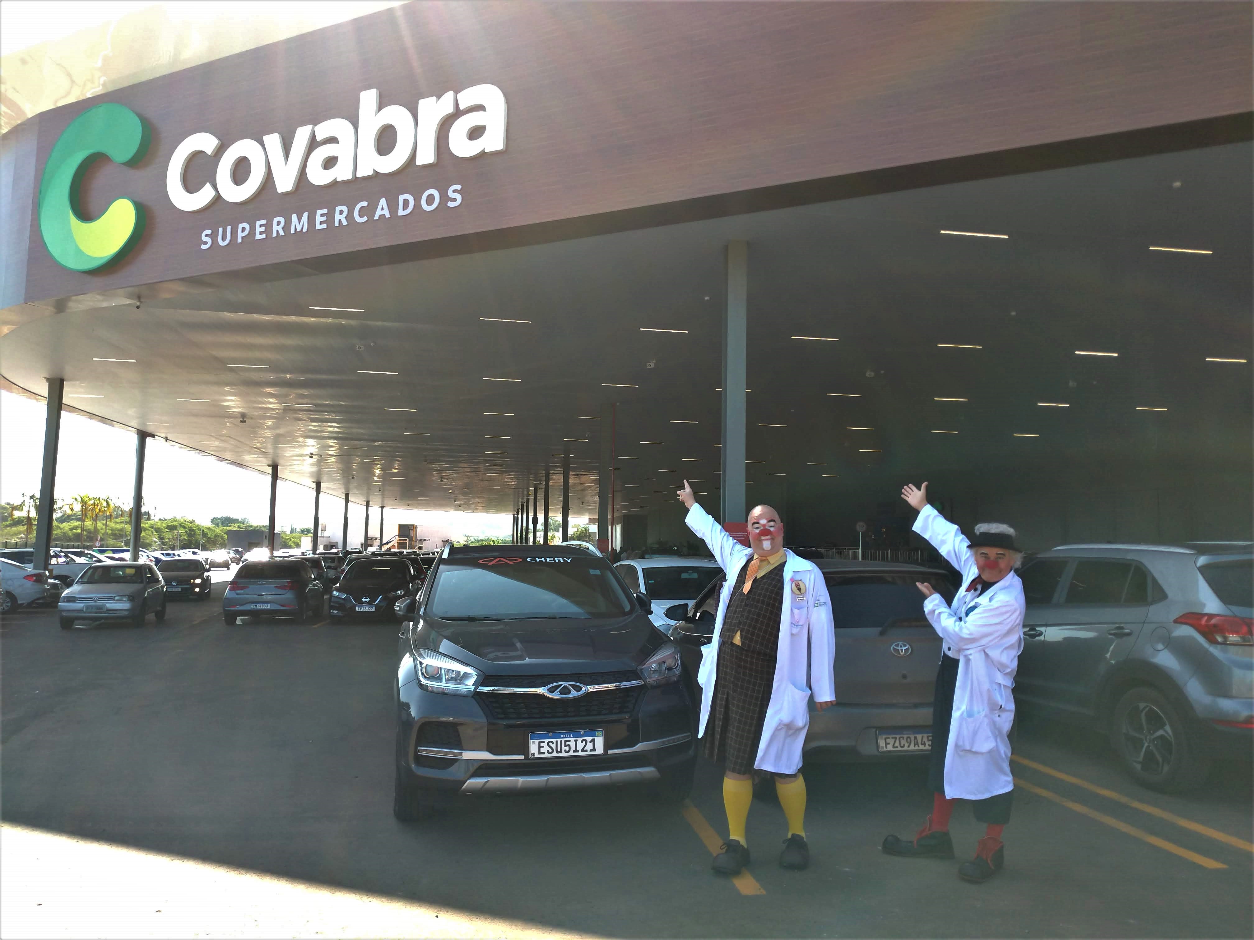 Cirurgiões Acerola e Gaguelho apontando para fachada e a marca Covabra Supermercados no estacionamento da nova loja em Limeira.